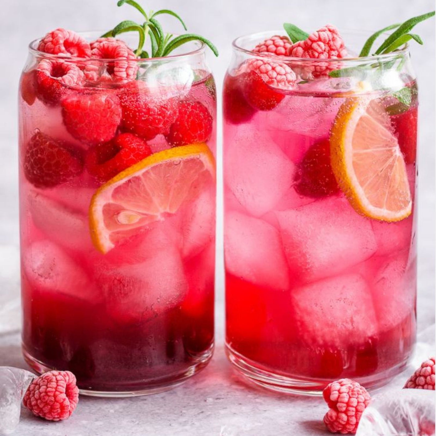 Two glasses of raspberry lemonade with ice and garnishes on a light background